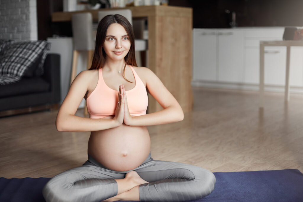 The Role of Yoga and Meditation in Enhancing Fertility 2 good looking pregnant caucasian female sportswear sitting yoga pose roll pad holding palms together namaste gesture smiling aside while relaxing thinking about baby scaled 1